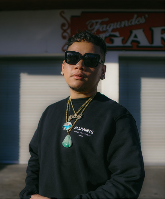 Dane Amar in sunglasses, a black sweatshirt and gold chains with a jade pendant, standing in late-afternoon sun outside a shop